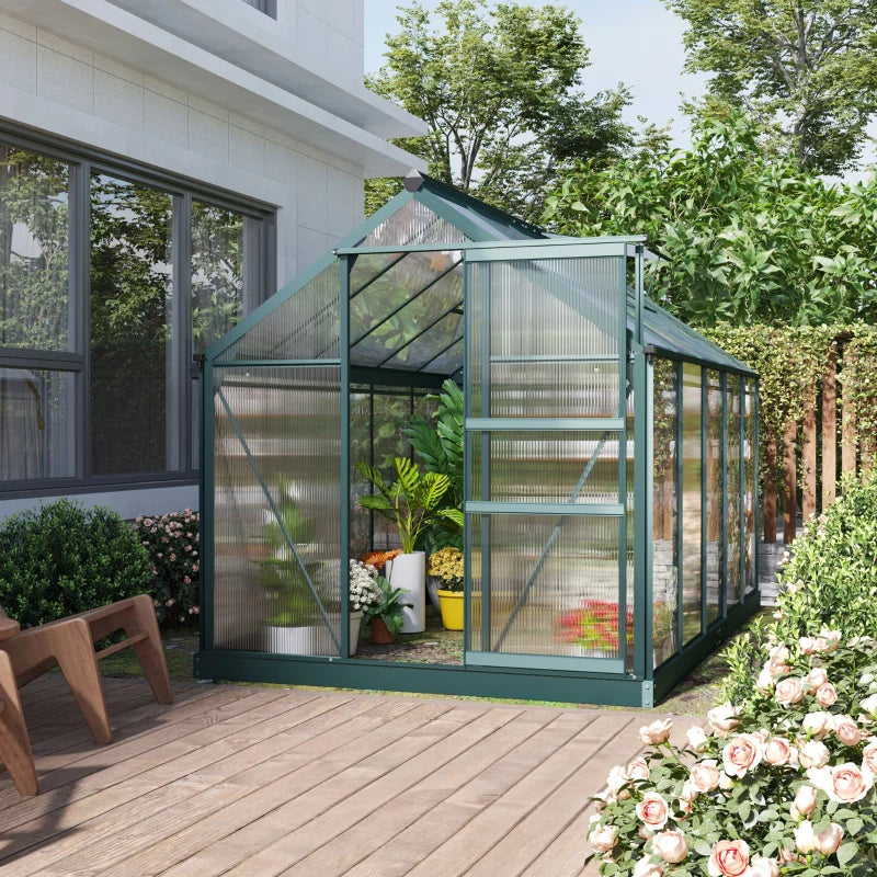 Green greenhouse attached to a house with plants inside, surrounded by flowers and trees.